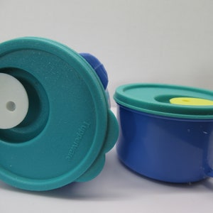 May include: Two blue and teal plastic containers with lids. The container on the left has a white lid with a small hole in the center. The container on the right has a handle and a yellow lid. The container on the left has the word "Tupperware" printed on the side.