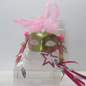 May include: A green and gold masquerade mask with pink feathers, silver leaves, and a pink star wand. The mask has a pearl accent and pink flowers.