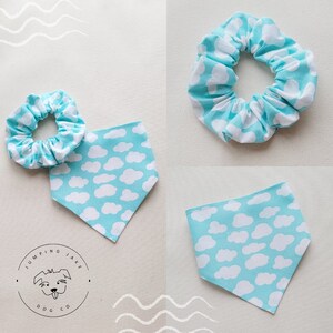 May include: A set of two light blue and white fabric accessories with a cloud pattern. The set includes a scrunchie and a bandana. The bandana is triangular shaped and the scrunchie is a circle. The scrunchie and bandana have a white cloud pattern on a light blue background. The text "Jumping Jake Dog Co." is printed on a white circle with a black outline.