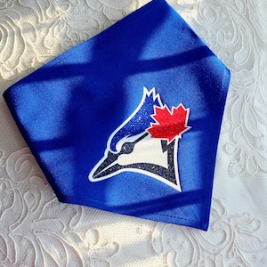 Può includere: Una bandana blu con il logo dei Toronto Blue Jays. Il logo include una testa di ghiandaia azzurra stilizzata, una foglia d'acero rossa e il nome della squadra. La bandana è realizzata in un tessuto liscio e lucido.