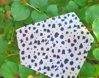 May include: A white bandana with a black dalmatian spot pattern. The bandana is square-shaped and is set against a backdrop of green foliage. The fabric appears to be soft and lightweight.