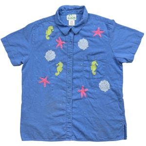 May include: Light blue short-sleeved shirt with embroidered seahorses, starfish, and seashells. The shirt has a button-down front, a collar, and a pocket on the left chest. The shirt is decorated with small, scattered embellishments.