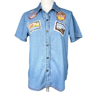 May include: Light blue denim short-sleeve shirt with embroidered patches. Patches include travel-themed designs and text. The shirt has a button-down front, a collar, and red stitching along the hem and collar.