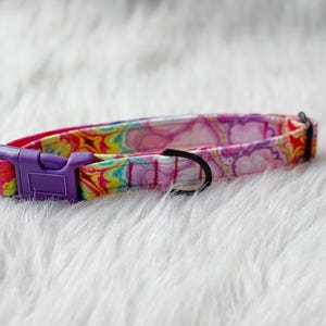 May include: A pet collar featuring a vibrant, multi-colored tie-dye pattern in shades of pink, yellow, and orange. The collar includes a purple plastic side-release buckle, a black metal D-ring for leash attachment, and a black plastic sliding adjuster.
