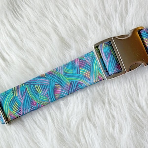May include: A fabric collar featuring a vibrant abstract pattern of intersecting teal, blue, purple, and yellow curved lines. The collar includes a polished silver-toned metal side-release buckle and a matching silver-toned metal rectangular adjustment slide.