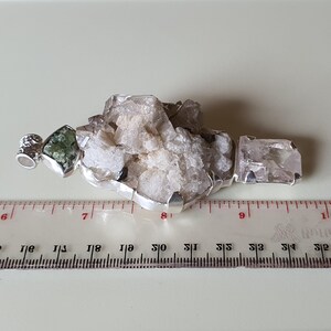 Puede incluir: Un colgante de plata con un grupo de cristales de cuarzo blancos y transparentes, una piedra verde y un cristal transparente. El colgante mide aproximadamente 7.6 cm de largo y se muestra sobre una superficie blanca.