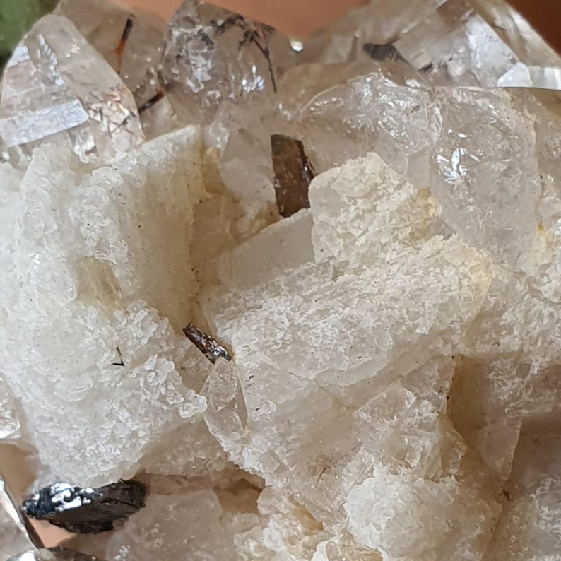 Puede incluir: Primer plano de un grupo de cristales de cuarzo. Los cristales son transparentes con inclusiones y se asientan sobre una matriz de formaciones minerales blancas y texturizadas. Peque&ntilde;os trozos oscuros de aspecto met&aacute;lico est&aacute;n dispersos por todo el grupo.