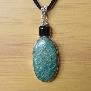 May include: A silver pendant necklace with a teal oval stone, a black square stone, and a small clear stone. The pendant hangs from a black cord. The oval stone has a striped pattern.