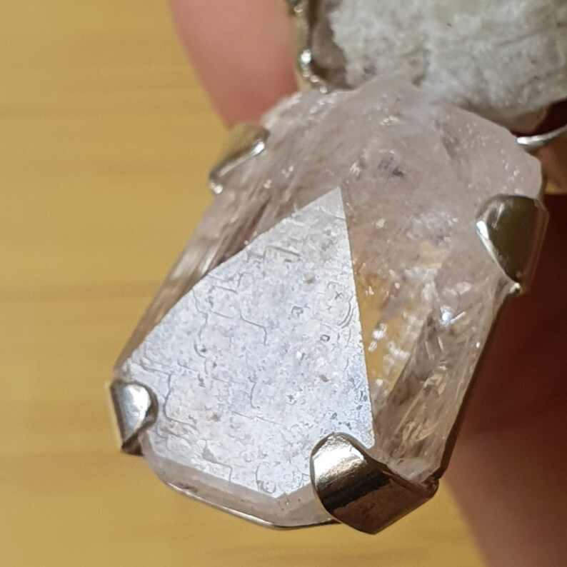 Puede incluir: Un colgante de cristal de cuarzo transparente con detalles met&aacute;licos plateados. El cristal es de un color claro y transl&uacute;cido con estructuras internas visibles. El engaste met&aacute;lico sujeta firmemente el cristal, mostrando su forma natural. El colgante es una joya &uacute;nica.