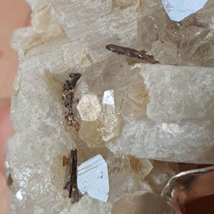 Puede incluir: Primer plano de un esp&eacute;cimen mineral con una matriz cristalina blanquecina. Contiene peque&ntilde;os cristales met&aacute;licos reflectantes, de color gris plateado y marr&oacute;n oscuro. La composici&oacute;n muestra formaciones geol&oacute;gicas naturales.