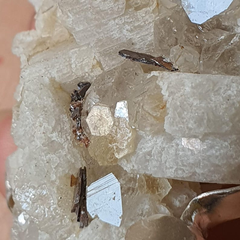Puede incluir: Primer plano de un esp&eacute;cimen mineral con una matriz cristalina blanquecina. Contiene peque&ntilde;os cristales met&aacute;licos reflectantes, de color gris plateado y marr&oacute;n oscuro. La composici&oacute;n muestra formaciones geol&oacute;gicas naturales.