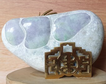 May include: A smooth, white jade stone with green and purple swirls, resting on a wooden surface. A gold-coloured metal ornament with a decorative design is in front of the stone.