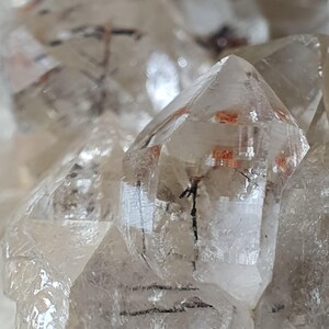 Puede incluir: Primer plano de cristales de cuarzo transparente con inclusiones internas. Los cristales tienen puntas afiladas y puntiagudas y exhiben una calidad transl&uacute;cida. Algunos cristales tienen inclusiones de color rojo-naranja y negro, lo que a&ntilde;ade inter&eacute;s visual. La disposici&oacute;n general sugiere un grupo de formaciones naturales.