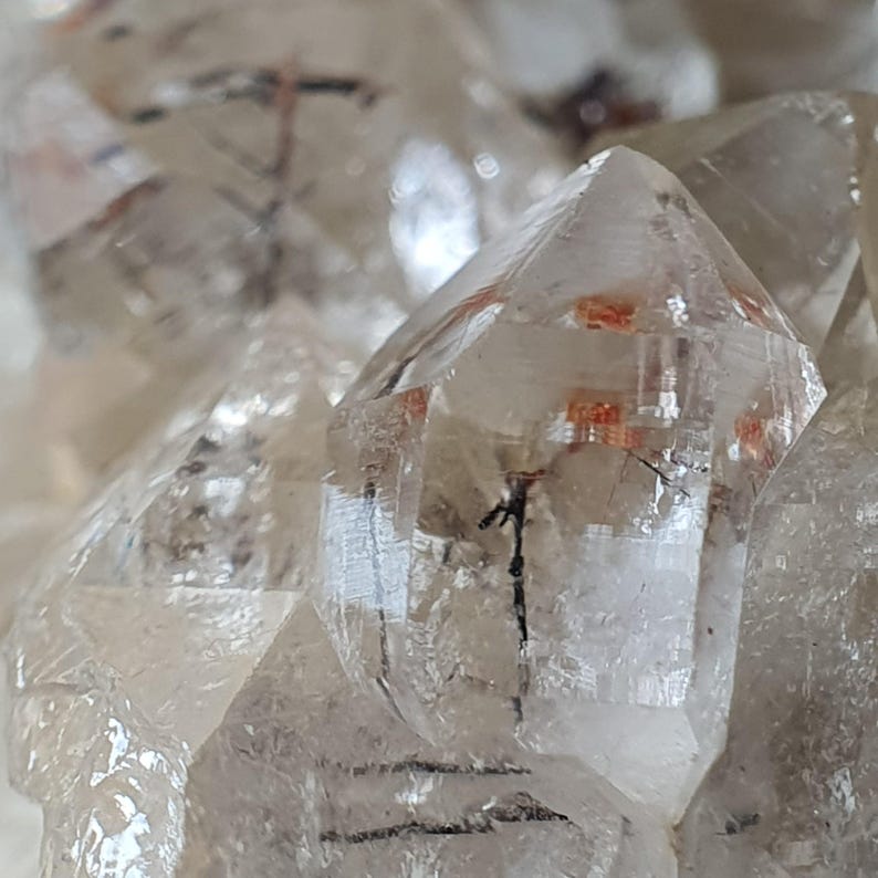 Puede incluir: Primer plano de cristales de cuarzo transparente con inclusiones internas. Los cristales tienen puntas afiladas y puntiagudas y exhiben una calidad transl&uacute;cida. Algunos cristales tienen inclusiones de color rojo-naranja y negro, lo que a&ntilde;ade inter&eacute;s visual. La disposici&oacute;n general sugiere un grupo de formaciones naturales.