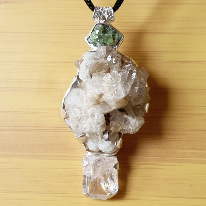 Puede incluir: Colgante &uacute;nico con un grupo de cristales blancos, una gema verde y un cristal claro rectangular. El colgante est&aacute; engastado en metal plateado y cuelga de un cord&oacute;n negro. Fondo de superficie de madera marr&oacute;n claro.
