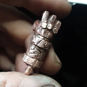 May include: A detailed copper-colored metal hand sculpture with a textured surface. The hand is closed into a fist with the fingers curled inwards.
