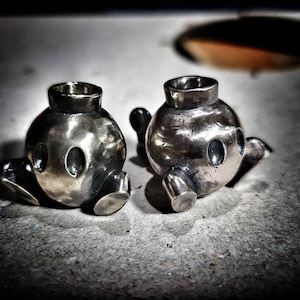 May include: Two silver-coloured, teapot-shaped metal objects with a rounded body, spout, and handle. Each teapot has a cylindrical opening at the top and two oval-shaped holes on the front. The teapots are placed on a textured, grey surface.