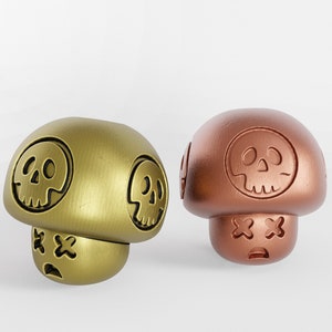 Pode incluir: Duas estatuetas de cogumelo de cerâmica com desenhos de caveiras e ossos cruzados. O cogumelo à esquerda é dourado e o da direita é cobre.