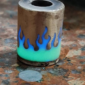 Op de afbeelding: Een zilveren kraal met een blauw en groen vlamontwerp. De kraal is cilindrisch en heeft een gat in het midden.