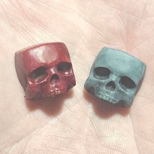 May include: Two small skull-shaped keycaps, one red and one grey, are shown on a hand. The keycaps are made of a smooth, textured material.