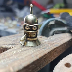 Op de afbeelding: Een klein, goudkleurig metalen beeldje van een robot met een enkele antenne op zijn hoofd. De robot heeft een tandenrij en een sigaar in zijn mond. Het beeldje staat op een houten oppervlak.