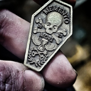 Può includere: Un ciondolo in metallo color argento a forma di bara con le parole "Memento Mori" e un design di teschio e ossa incrociate.