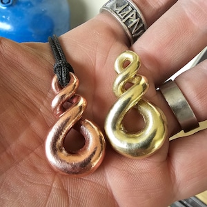 May include: Two infinity symbol pendants, one copper and one gold, on a black cord. The pendants are on a hand wearing two rings.