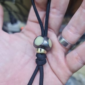Op de afbeelding: Een zwart koord met een paddenstoelvormige hanger. De hanger heeft een zilverkleurige dop met een lichtgroene bovenkant en een goudkleurige basis. Het koord is gemaakt van een geweven zwart koord.