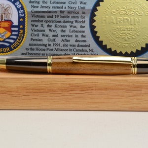 May include: A handcrafted pen with a gold-colored tip and accents, and a dark wood barrel. The pen rests on a wooden base, with a document and seal in the background. The pen is a writing instrument.