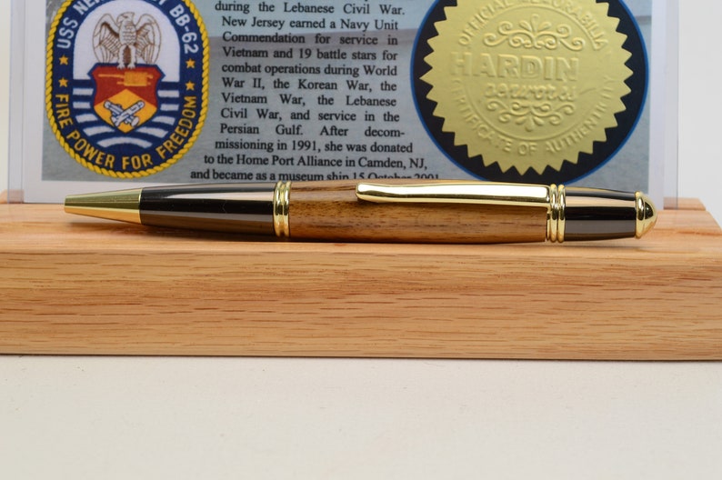 May include: A handcrafted pen with a gold-colored tip and accents, and a dark wood barrel. The pen rests on a wooden base, with a document and seal in the background. The pen is a writing instrument.