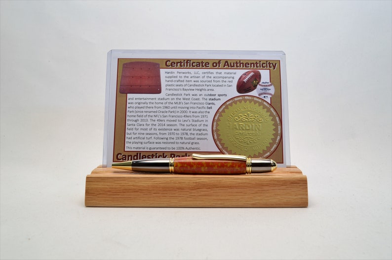 May include: A handcrafted pen with a red and orange speckled barrel, gold accents, and a silver tip, displayed on a wooden stand. A certificate of authenticity is visible behind the pen, with text about Candlestick Park.
