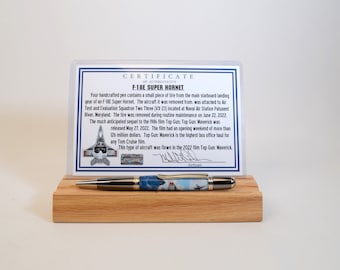 May include: A handcrafted pen with a blue and white design, displayed on a wooden stand. The pen is accompanied by a certificate of authenticity for an F-18E Super Hornet. The certificate includes text and an image of the aircraft.