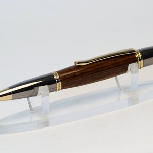 May include: A handcrafted pen with a dark wood barrel, accented with gold and silver-toned metal. The pen features a gold-colored tip and clip. It is displayed on a clear acrylic stand, showcasing its elegant design.