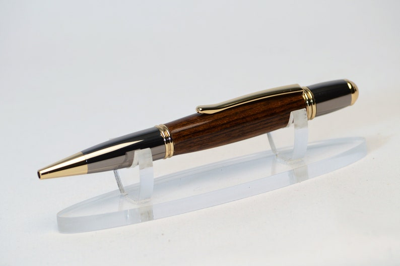 May include: A handcrafted pen with a dark wood barrel, accented with gold and silver-toned metal. The pen features a gold-colored tip and clip. It is displayed on a clear acrylic stand, showcasing its elegant design.