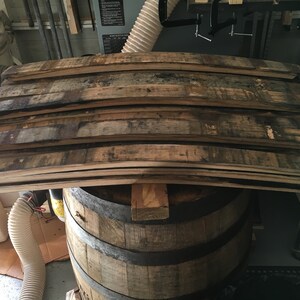 K&ouml;nnte beinhalten: Ein Stapel gebogener Holzdauben aus recycelten F&auml;ssern, die Farbschattierungen von hell- bis dunkelbraun aufweisen. Die Dauben liegen auf einem teilweise montierten Holzfass mit Metallb&auml;ndern. Das Bild deutet auf ein Holzbearbeitungs- oder Upcycling-Projekt hin.