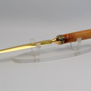 May include: A gold-colored letter opener with a wooden handle and a black tip. The handle has a decorative gold band. The letter opener is displayed on a clear acrylic stand. The blade is long and slender.