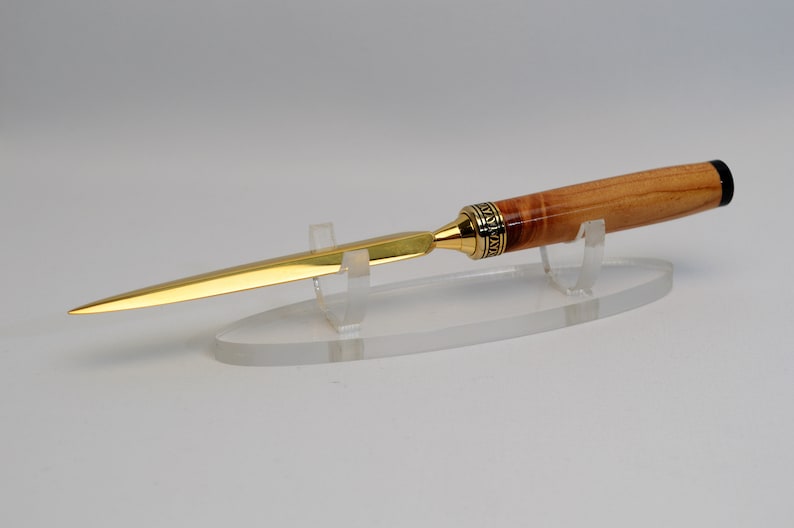 May include: A gold-colored letter opener with a wooden handle and a black tip. The handle has a decorative gold band. The letter opener is displayed on a clear acrylic stand. The blade is long and slender.