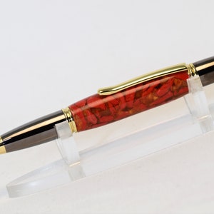 May include: A handcrafted pen with a red and gold patterned barrel, black accents, and gold-colored trim. The pen is displayed on a clear acrylic stand, showcasing its elegant design. The pen's tip and end cap are also gold-colored.
