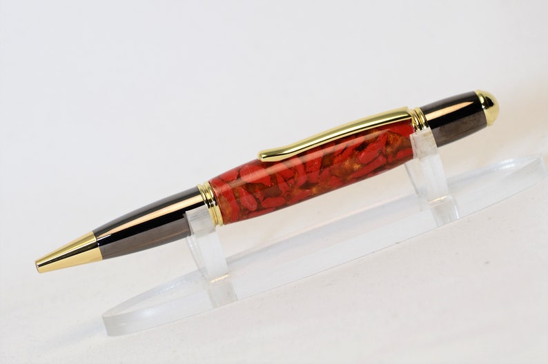 May include: A handcrafted pen with a red and gold patterned barrel, black accents, and gold-colored trim. The pen is displayed on a clear acrylic stand, showcasing its elegant design. The pen's tip and end cap are also gold-colored.