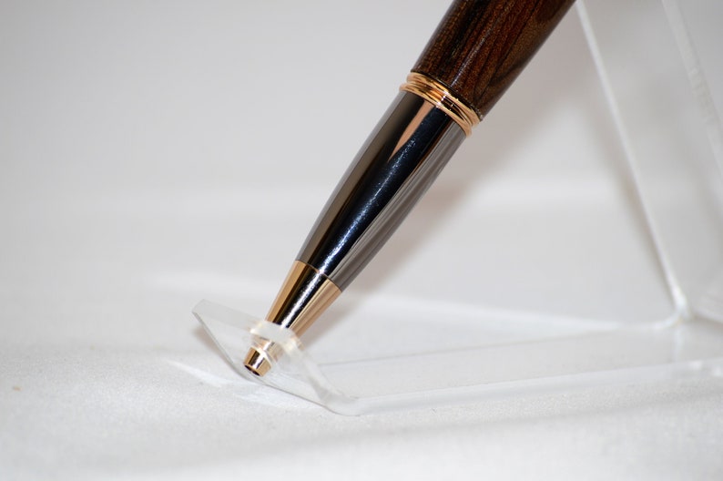 Puede incluir: Primer plano de un bol&iacute;grafo con un cuerpo de madera oscura, una banda dorada y un cuerpo gris met&aacute;lico. La punta del bol&iacute;grafo es dorada. El bol&iacute;grafo descansa sobre un soporte de pl&aacute;stico transparente sobre un fondo blanco.