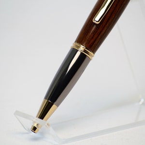 May include: A close-up of a wooden pen with gold and black accents. The pen has a dark brown, wood-grain barrel, a black metal center section, and gold-colored trim. The pen is displayed on a clear acrylic stand against a white background.
