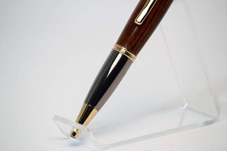 May include: A close-up of a wooden pen with gold and black accents. The pen has a dark brown, wood-grain barrel, a black metal center section, and gold-colored trim. The pen is displayed on a clear acrylic stand against a white background.