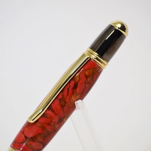 May include: A close-up of a pen with a red and gold patterned barrel. The pen has gold accents, including the clip and the top of the cap. The cap is black with a gold tip.
