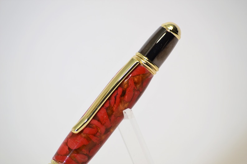May include: A close-up of a pen with a red and gold patterned barrel. The pen has gold accents, including the clip and the top of the cap. The cap is black with a gold tip.