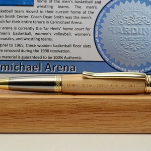 Peut inclure: Un stylo en bois avec des d&eacute;tails dor&eacute;s et argent&eacute;s repose sur un socle en bois. Le stylo a un corps en bois clair et une pointe dor&eacute;e. Un panneau bleu et blanc avec du texte et un sceau est en arri&egrave;re-plan. Le texte indique "Carmichael Arena."