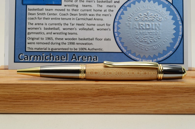 Peut inclure: Un stylo en bois avec des d&eacute;tails dor&eacute;s et argent&eacute;s repose sur un socle en bois. Le stylo a un corps en bois clair et une pointe dor&eacute;e. Un panneau bleu et blanc avec du texte et un sceau est en arri&egrave;re-plan. Le texte indique "Carmichael Arena."