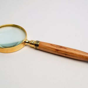 May include: A magnifying glass with a gold-colored rim and handle accents. The handle is made of light-colored wood. The lens is clear, and the handle has a dark, black tip. The magnifying glass is a tool for reading or examining small objects.