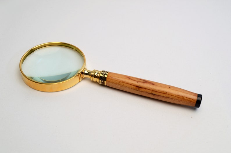 May include: A magnifying glass with a gold-colored rim and handle accents. The handle is made of light-colored wood. The lens is clear, and the handle has a dark, black tip. The magnifying glass is a tool for reading or examining small objects.