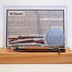 Könnte beinhalten: Ein Holzstift mit einer Geschossspitze und Metallakzenten, präsentiert auf einem Holzständer. Dahinter befindet sich ein gerahmtes Dokument über das M1 Garand Gewehr, mit einem Echtheitszertifikat-Siegel.