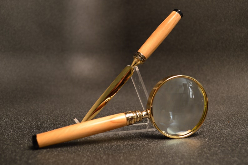 May include: A letter opener and magnifying glass. The letter opener has a gold blade and a light wood handle with a black tip. The magnifying glass has a gold frame and a light wood handle.